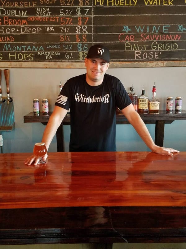Mike See posing behind the Witchdoctor Brewing Bar with the menu and some of their specialty liquors